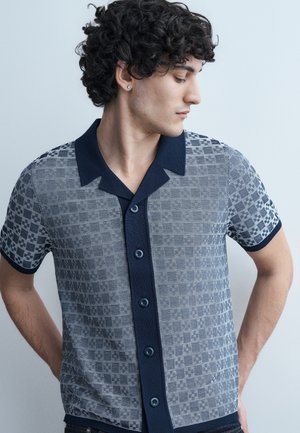 Άνδρας με σγουρά μαλλιά, φοράει πουκάμισο με κοντά μανίκια και γιακά σε ναυτικό μπλε, με μοτίβο μπλε και λευκού υφάσματος, κοιτάζει προς τα αριστερά του.