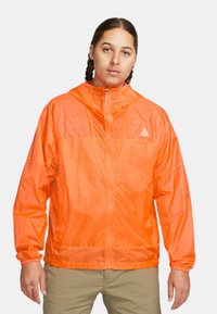 Orange lättviktsjacka med dragkedja framtill och huva. Har elastiska manschetter och en diskret triangellogo på bröstet. Slät textur.