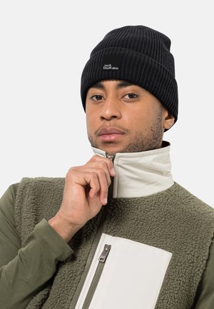 Black ribbed knit beanie with stitched logo, paired with a green fleece jacket featuring a white zippered pocket and high collar.