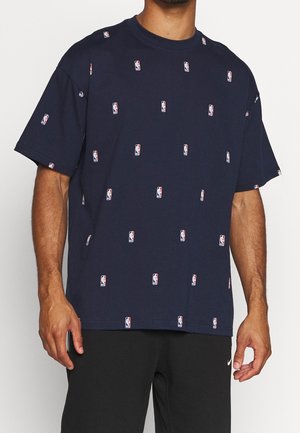 Homme portant un t-shirt bleu marine orné de petits logos NBA et un pantalon Nike noir, debout devant un fond uni.