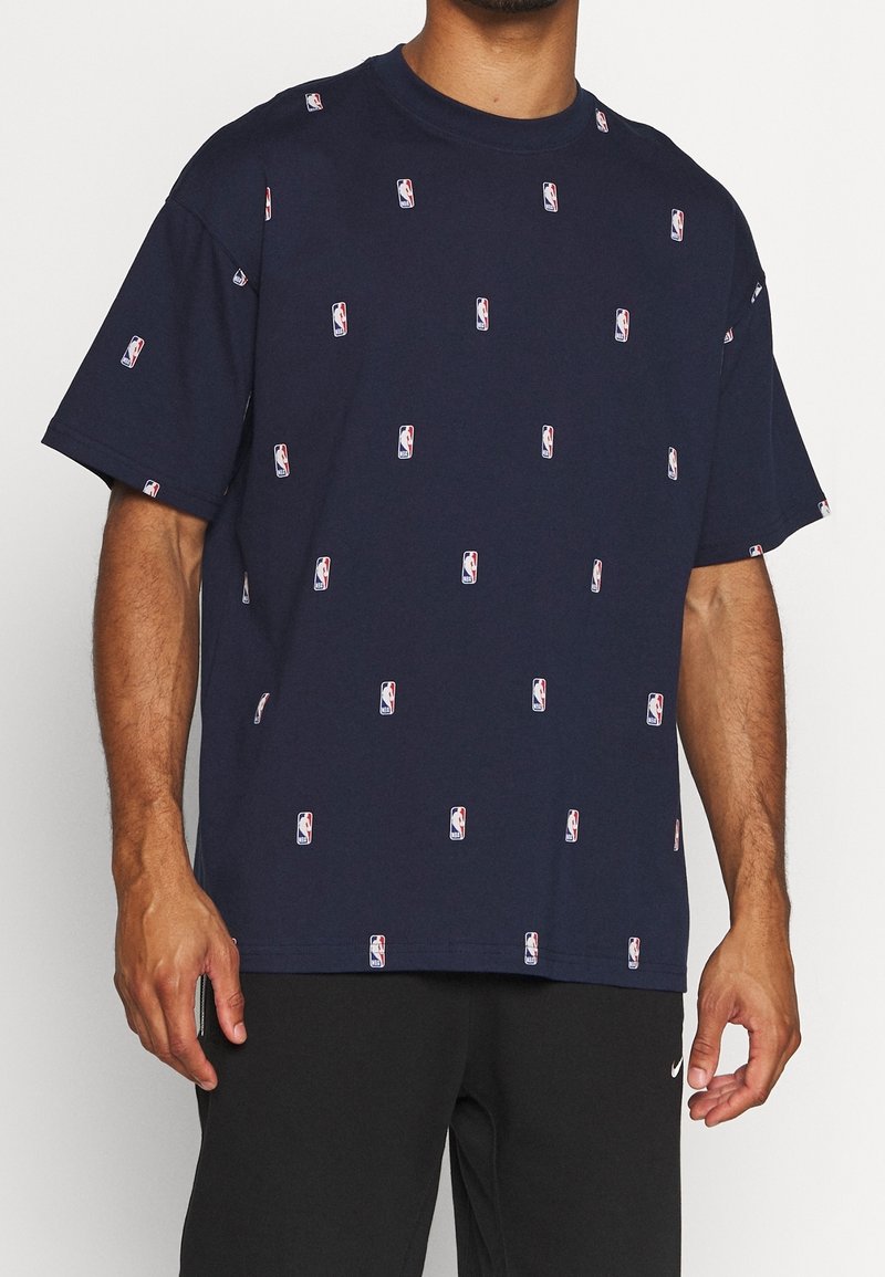 Homme portant un t-shirt bleu marine orné de petits logos NBA et un pantalon Nike noir, debout devant un fond uni.