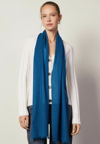 Écharpe bleue en matériau doux, présentant une texture lisse et des bords effilochés. Portée avec un cardigan blanc, mettant en valeur sa couleur vive.