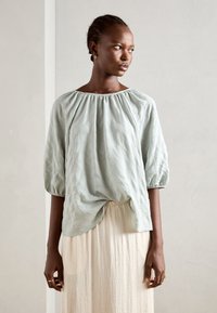 Lys grønn bluse med samlet halslinje og puffermer, laget av teksturert stoff. Brukt over et flytende kremfarget skjørt.