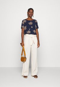 Blusa de manga curta com estampado floral marinho, combinada com calças largas em creme com um cinto amarrado. Acessorizada com uma bolsa de ombro castanha.