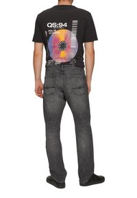 Schwarzes Grafik-T-Shirt mit einem runden farbigen Vinyl-Design auf dem Rücken, kombiniert mit dunkelgrauen Jeans und schwarzen Schuhen.
