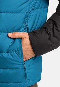 Giacca isolante turchese con maniche nere, dotata di una tasca con zip e di una texture trapuntata per garantire calore e comfort.