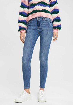 Kvinde iført lyseblå skinny jeans, en stribet striktrøje i pink, lilla, grøn og hvid, og hvide sneakers, stående foran en ensfarvet baggrund.