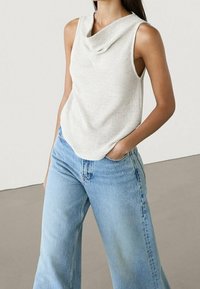 Woman wearing sleeveless white textured top with draped neckline and light blue high-waisted wide-leg jeans, standing with one hand in pocket.
