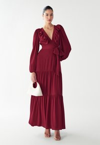 Dlhé burgundské šaty s volánmi na v�ýstrihu a rukávoch, s viazaným pásom a stupňovaným lemom; v kombinácii s bielou kabelkou.