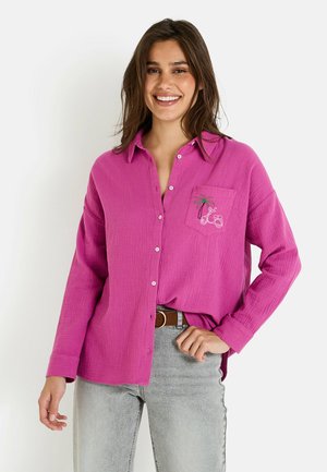 Chemise rose à boutons avec une finition texturée, dotée d'une poche poitrine avec une broderie de palmier et de scooter. Portée avec un jean gris.