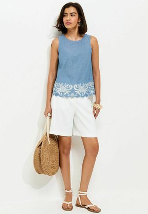 Vrouw draagt een mouwloos blauw topje met witte bloemrijke borduursels, witte korte broek, witte sandalen, draagt een ronde strozak en staat tegen een witte achtergrond.