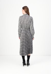 Schwarz-weiß gemustertes Kleid mit langen Ärmeln, gefälteltem Design, tailliertem Bund und knielanger Länge. Getragen mit schwarzen Schuhen mit Absätzen und Socken.