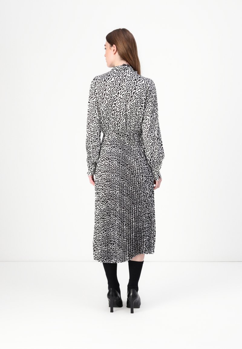 Schwarz-weiß gemustertes Kleid mit langen Ärmeln, gefälteltem Design, tailliertem Bund und knielanger Länge. Getragen mit schwarzen Schuhen mit Absätzen und Socken.