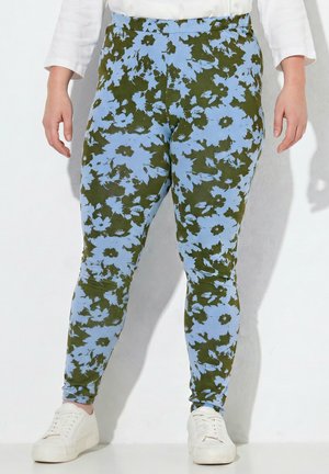 Personne portant des leggings moulants à motif floral bleu et vert, des baskets blanches et un haut blanc à manches longues, debout contre un mur.