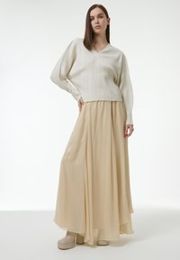 Frau trägt einen cremefarbenen, gerippten V-Ausschnitt-Pullover, einen fließenden, beigen Maxirock und beige Plateaustiefel, steht vor einem weißen Hintergrund.