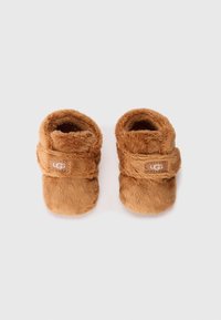 Chaussons pour bébés en peluche brune avec une bande velcro et le logo UGG, présentant une texture douce et moelleuse ainsi qu'une forme arrondie, conçus pour le confort.