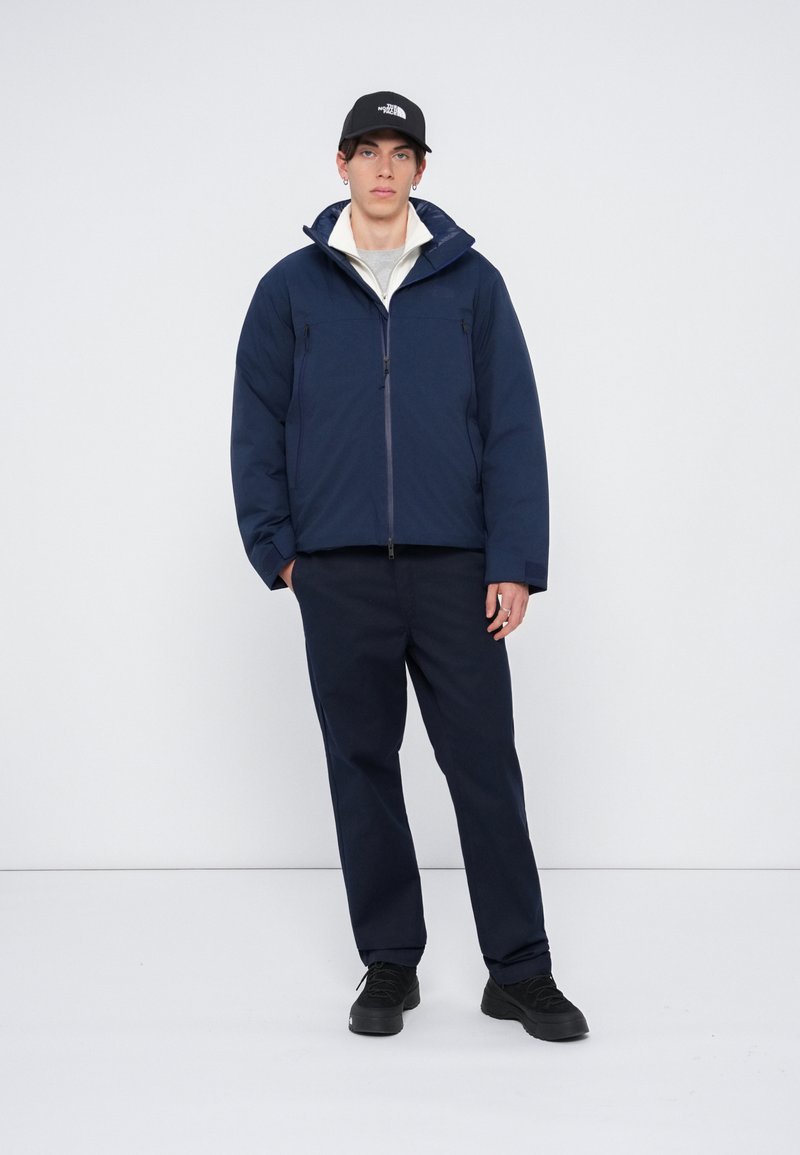 Veste matelassée marine avec capuche, deux poches zippées et une texture unie. Associée à un pantalon marine assorti et des baskets noires.