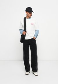 Jeune homme portant une casquette noire, une chemise blanche à manches courtes sur une chemise bleue à manches longues, un pantalon noir, des baskets blanches et portant un sac à bandoulière noir.