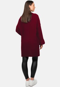 Cardigan long en maille couleur bordeaux, coupe ample, texture côtelée, avec fentes latérales, porté avec des leggings noirs en simili cuir et des baskets blanches.