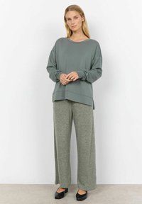 Groen langemouwen top met een ronde hals en zijsplitten, gecombineerd met lichtgrijze wijde broek. Zwarte gesloten schoenen maken de outfit compleet.