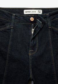 Donkerblauwe denim jeans met een ritssluiting, koperen knoop en contrasterende gele stiksels. Label toont maat W 27 L 32. Voorkruiden zichtbaar.