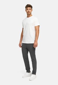 Ung mann står, iført en hvit t-skjorte, mørkegrå joggebukser og hvite joggesko mot en ensfarget lysegrå bakgrunn.