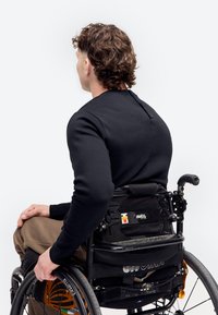 Homme aux cheveux bouclés portant une chemise noire à manches longues, assis dans un fauteuil roulant noir, dos tourné vers un fond blanc.