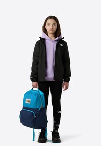 Blå og marineblå ryggsekk med kontrasterende glidelåser, båret av en ungdom i svart jakke over en lavendelfarget hettegenser og svarte leggings.