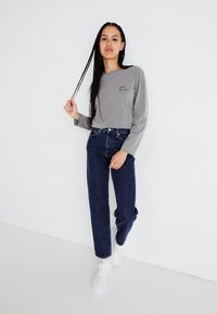 Grå langermet topp med lite logo på brystet, kombinert med mørkeblå rettbente jeans og hvite joggesko. Minimalistisk design.