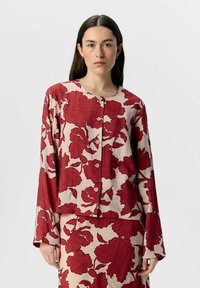 Rød jakke med blomstermotiv på en beige baggrund, lavet af tekstureret stof. Har rund halsudskæring og knaplukning foran.