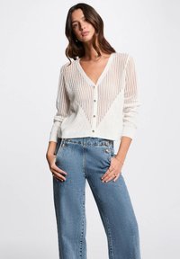 Cardigan bianco a maglia con scollo a V, caratterizzato da un motivo testurizzato e bottoni sul davanti. Abbinato a jeans in denim blu a vita alta.