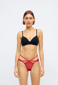 Μαύρο στερεωμένο σουτιέν με λεπτές τιράντες, συνδυασμένο με κόκκινο λαμέ στρινγκ με διασταυρούμενες τιράντες και πάνελ από διαφαινές ύφασμα, αναδεικνύοντας την περίπλοκη λεπτομέρεια.