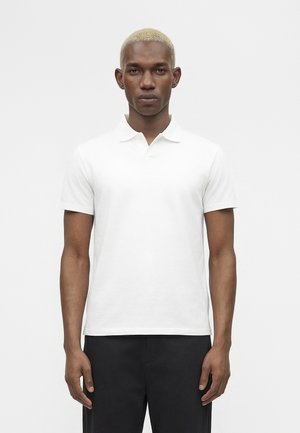 Moški s kratkimi blond lasmi, oblečen v enostavno belo polo majico s kratkimi rokavi in črne hlače, stoji pred belim ozadjem.