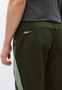 Calças verde-oliva com acabamento texturizado, com uma faixa lateral verde-claro e uma etiqueta preta CLIMAWARM no bolso traseiro.