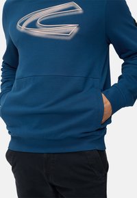 Blå sweatshirt med stort hvidt logo foran på brystet. Har ribbet kant forneden og ved ærmerne samt sidelommer for funktionalitet.