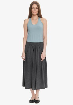 Kobieta stojąca, ubrana w jasnoniebieski top bez rękawów, ciemnoszare długie spódnice z kieszeniami oraz czarne balerinki na białym tle.