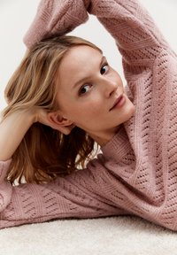 Helles rosa gestricktes Sweatshirt mit strukturierten, offenen Mustern. Das Model hat lange, glatte Haare und posiert mit Händen hinter dem Kopf vor einem neutralen Hintergrund.