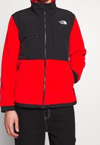 Mężczyzna w czarnej i czerwonej polarowej kurtce z zamkiem, z logo The North Face na piersi, z jedną ręką w kieszeni, na jednolitym tle.