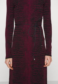 Femme portant une robe noire à manches longues avec un motif en grille irrégulier rose foncé et une fine ceinture noire nouée à la taille, se tenant devant un fond blanc.