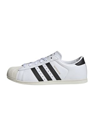 Baskets basses blanches avec des bandes noires, un bout texturé, des lacets blancs et le texte "Superstar" sur le côté.
