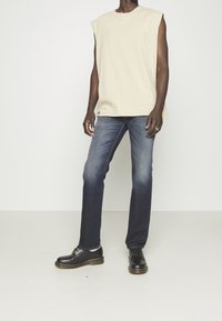 Camisa sin mangas beige, jeans azul oscuro, zapatos negros con costuras amarillas. Jeans de corte recto con un ligero desgaste, diseño simple en toda la prenda.