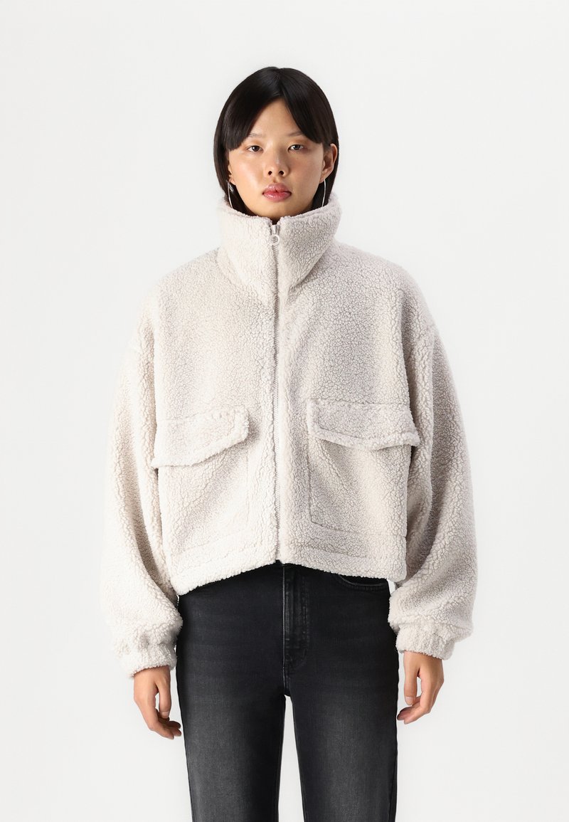Lyhennetty kermaisen värinen fleece-takki, jossa on korkea kaulus, kaksi etutaskua ja joustavat hihansuut. Pehmeä rakenne ja rento istuvuus.