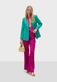 Blazer texturizado en teal sobre una camisa de satén rosa, combinado con pantalones acampanados rosas. Los accesorios incluyen un bolso dorado y sandalias de tacón.