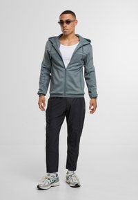 Světle šedá mikina na zip s kapucí a texturovanými panely, doplněná o černé zúžené kalhoty. Model má na sobě sportovní boty a sluneční brýle.