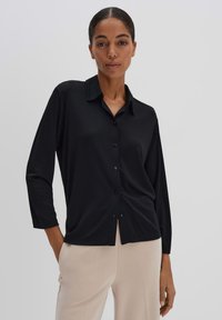 Blouse noire à manches longues avec boutons sur le devant et col. Tissu doux, coupe décontractée et légère fronce à l'ourlet. Portée avec un pantalon clair.