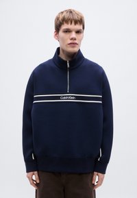 Μπλε ναυτικό φούτερ με ψηλό γιακά, κλείσιμο με φερμουάρ στη μέση και δύο οριζόντιες λευκές ρίγες. Διαθέτει το λογότυπο Calvin Klein στο στήθος.