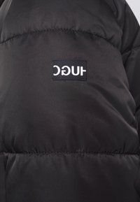 Chaqueta acolchada negra con textura de acolchado. Presenta una etiqueta de logo prominente. El material parece suave y resistente al agua.