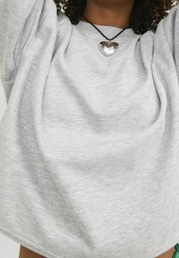 Felpa grigia chiara a maniche corte con vestibilità morbida e tessuto morbido e testurizzato. Presenta un ciondolo a forma di cuore argento su una collana nera.