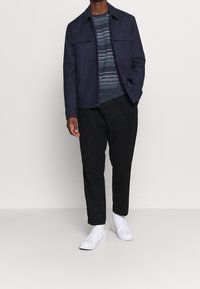 Námořnická bunda se dvěma předními kapsami, přes modré pruhované svetr, v kombinaci s černými kalhotami a bílými teniskami.