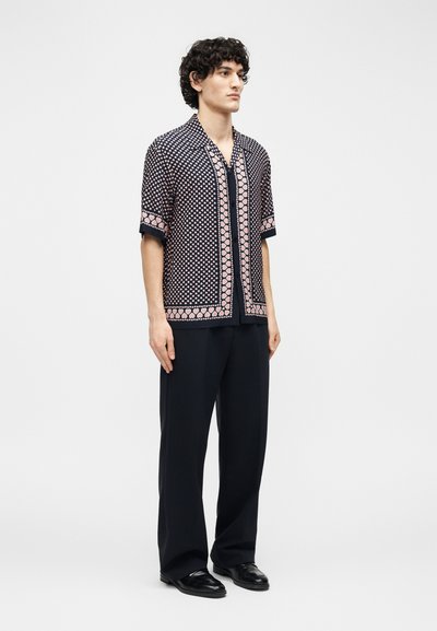 Νεαρός άντρας όρθιος, φοράει κοντομάνικο μπλε πουκάμισο με ροζ γεωμετρικά μοτίβα, μαύρο παντελόνι φαρδιάς γραμμής και μαύρα παπούτσια.